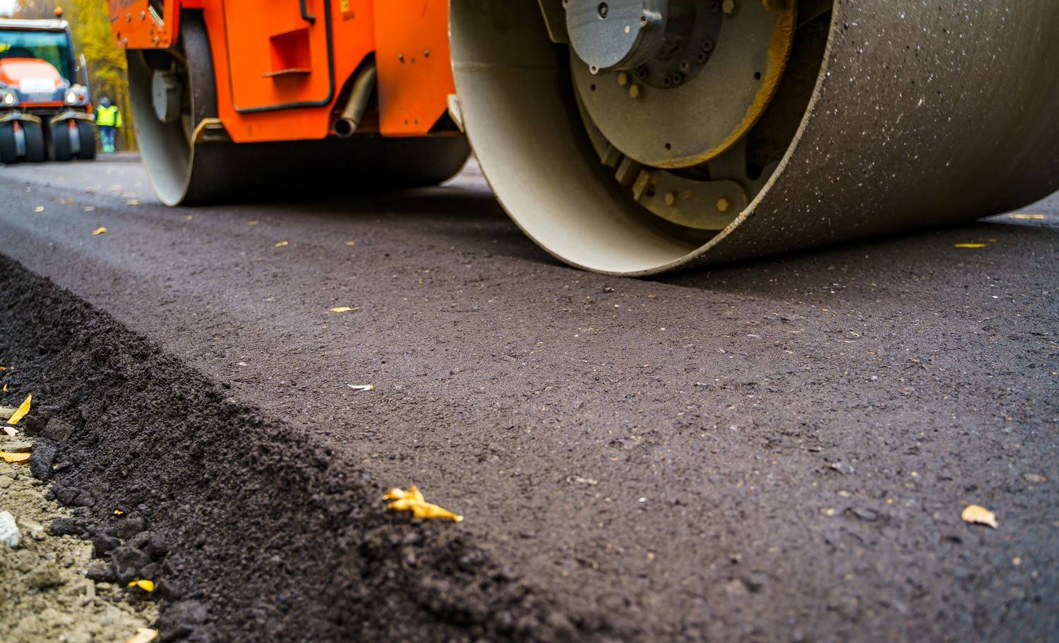 Worker in high-visibility gear spreading fresh hot-mix asphalt during a road paving project.